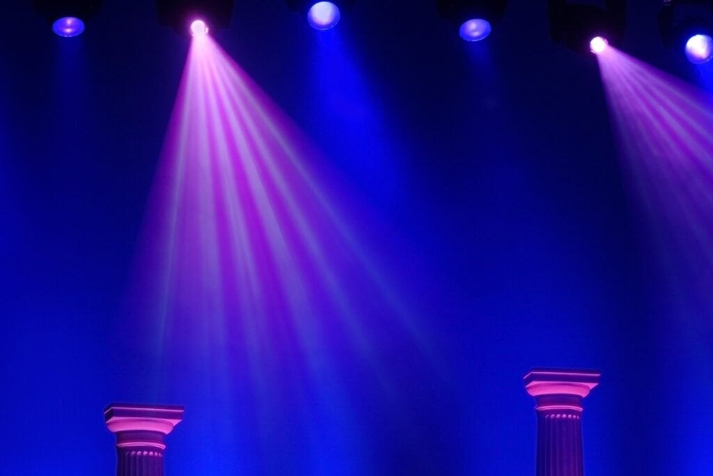 Approximately six blue and violet stage spot lights shinning from above the stage. The lights are hanging and the frame supporting them is slightly visible. The stage lights are pointed downward toward two Greek columns. There is fog in the air creating richer tones from the lights.