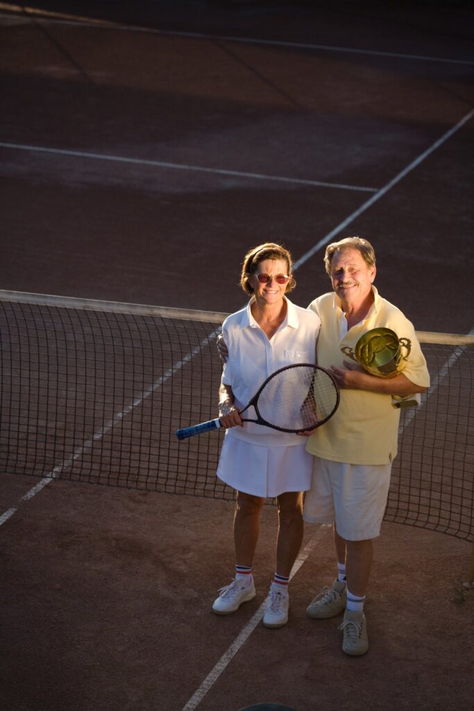 Active senior couple is posing on the tennis court with tennis racket and cup in hand. Outdoor, sunlight.